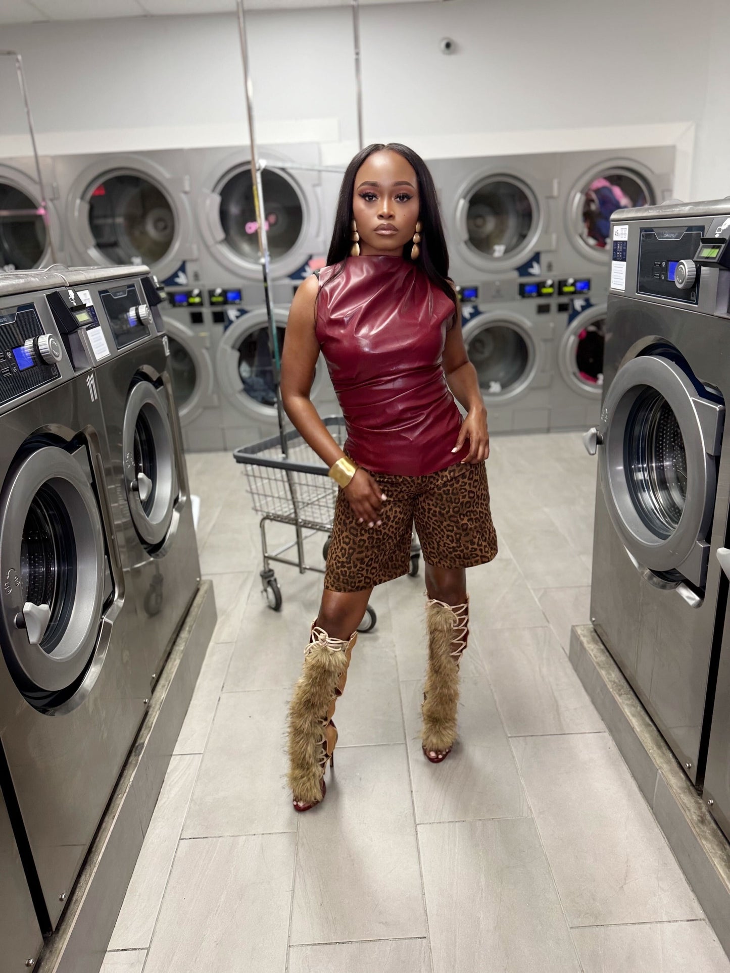 Woman posing in a laundromat with washing machines in the background in a red colored leather top and leopard print jean shorts
