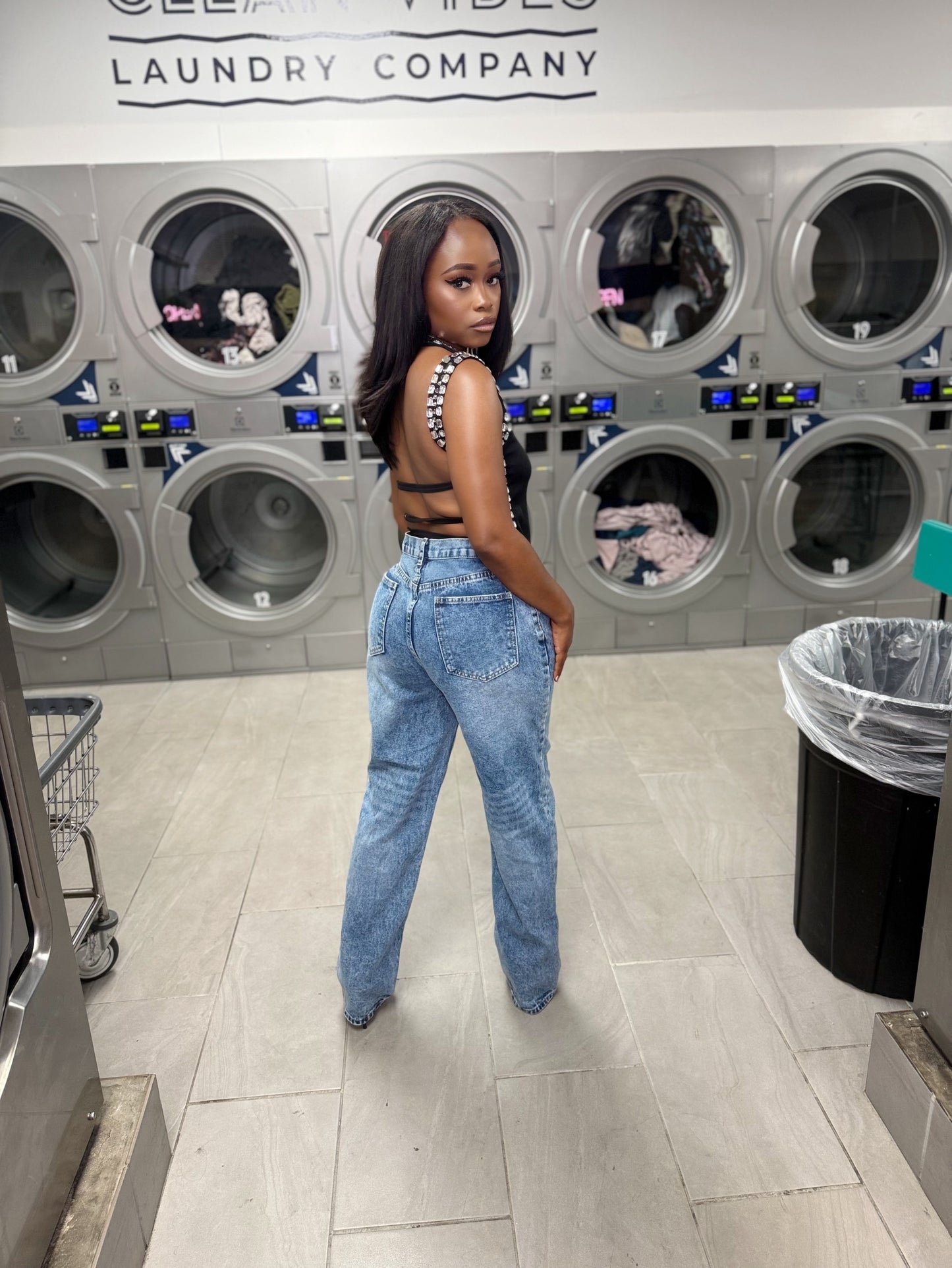 Woman standing in a laundromat with washing machines in the background in a black open back embellished top