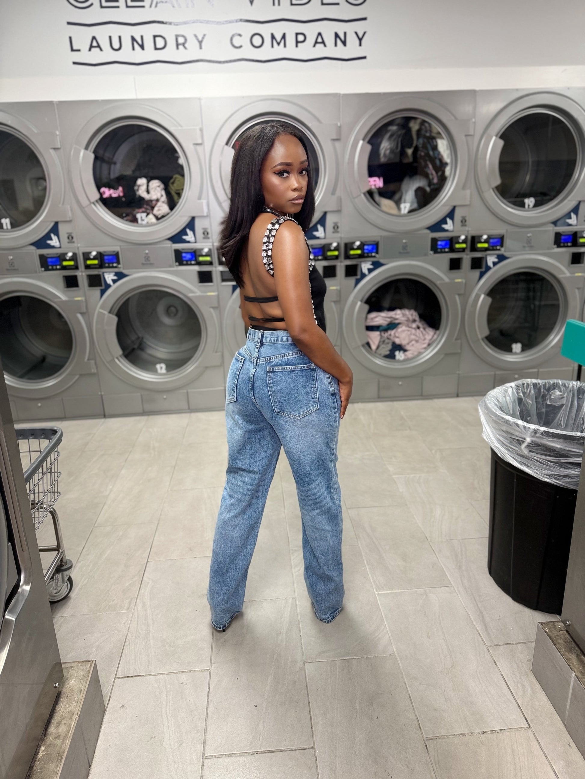 Woman standing in a laundromat with washing machines in the background in a black open back embellished top
