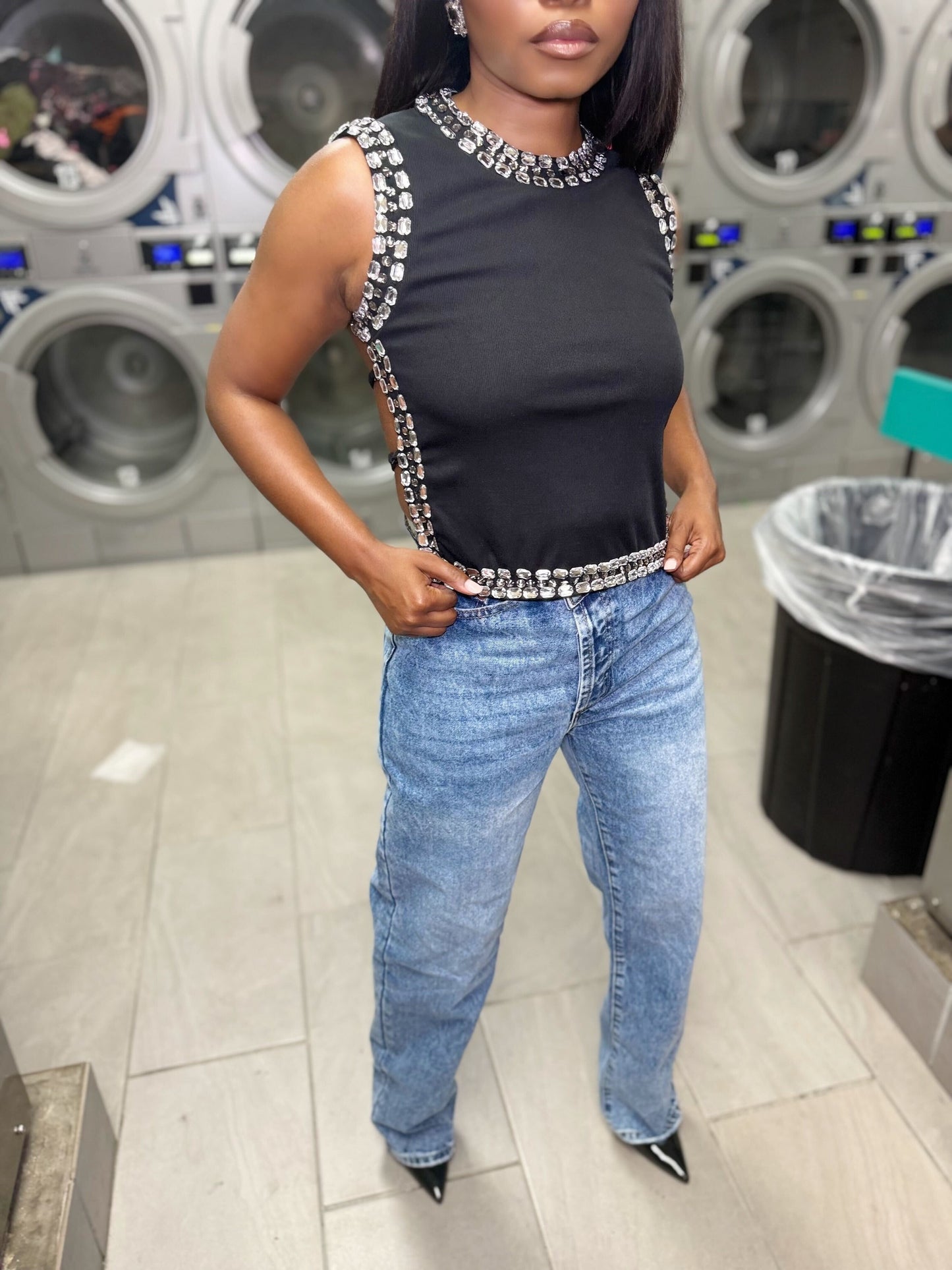 Person wearing a black top with decorative straps and blue jeans in a laundromat.